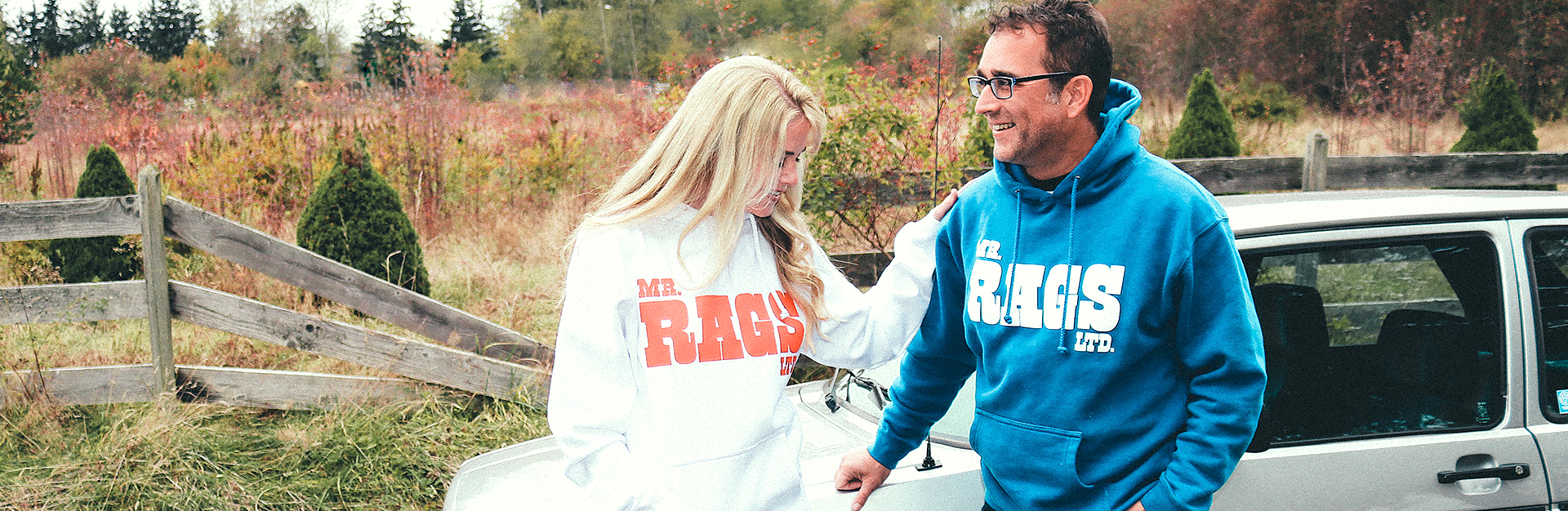 Two people wearing white and blue Mr. Rags hoodies beside a vintage car